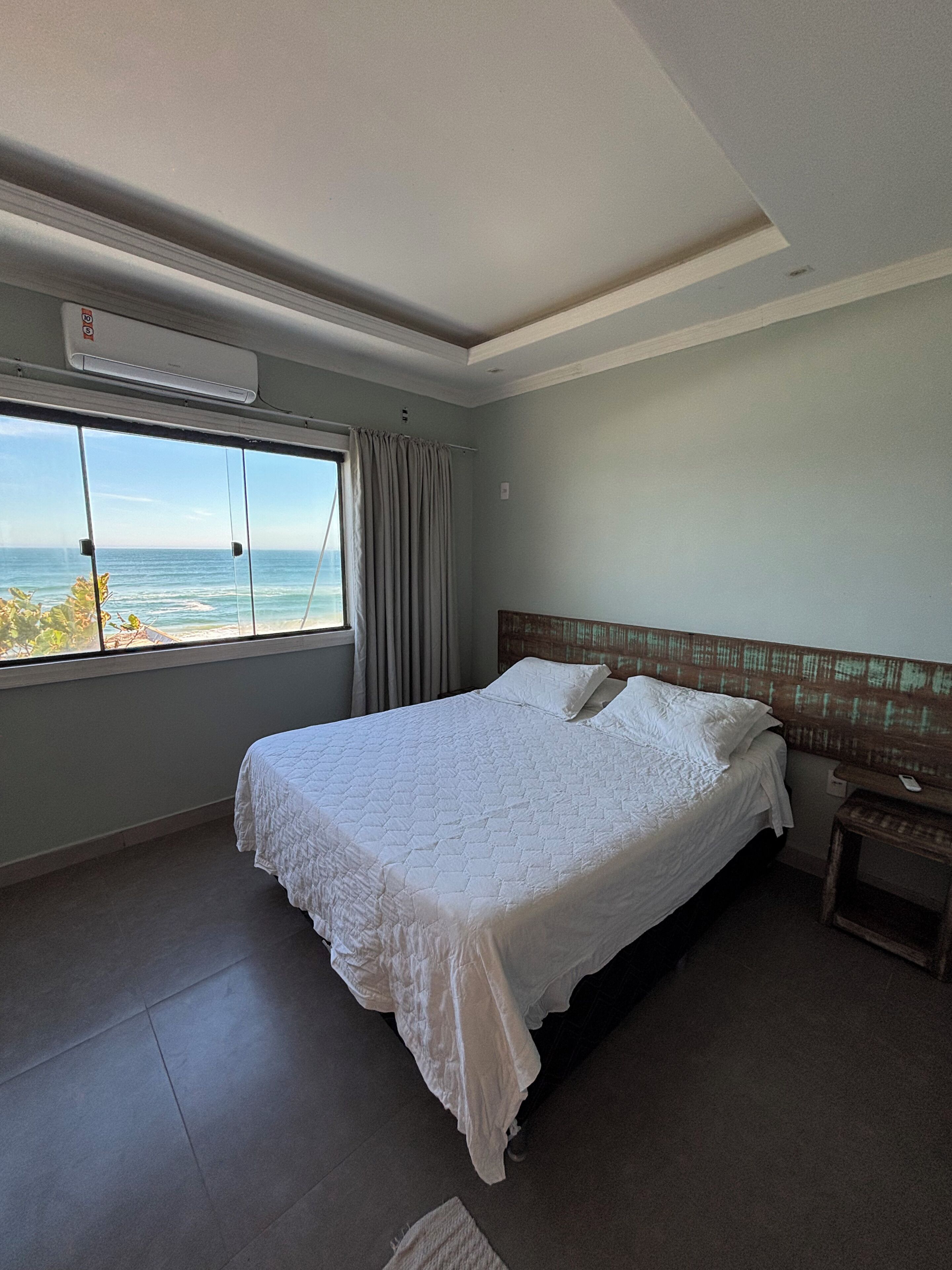 A hotel room with a bed, a nightstand, a window with a view of the ocean, and an air conditioning unit.