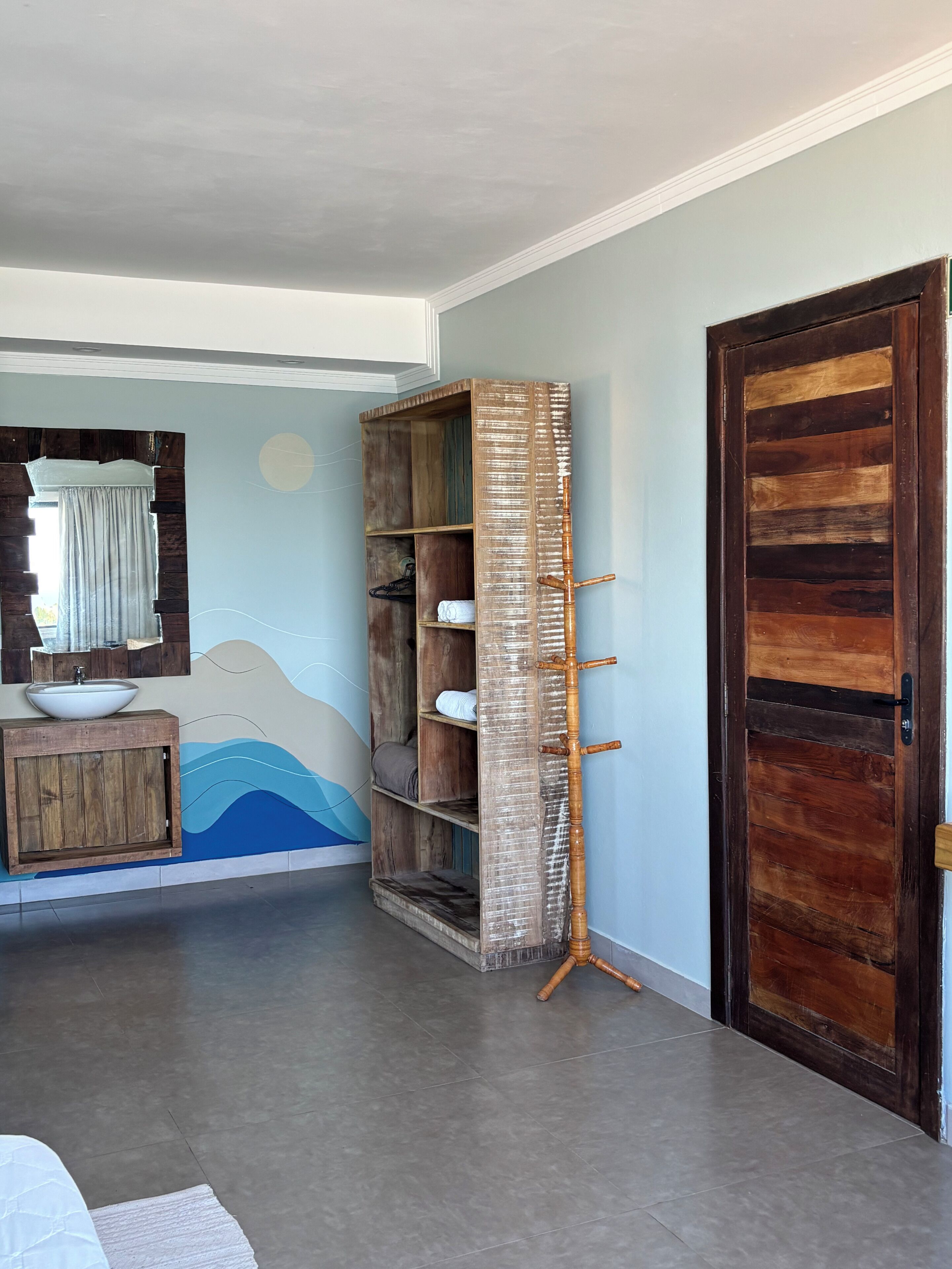 A room with a wooden cabinet, a wall-mounted mirror, a large wooden door, and a floor-to-ceiling wooden shelf unit.