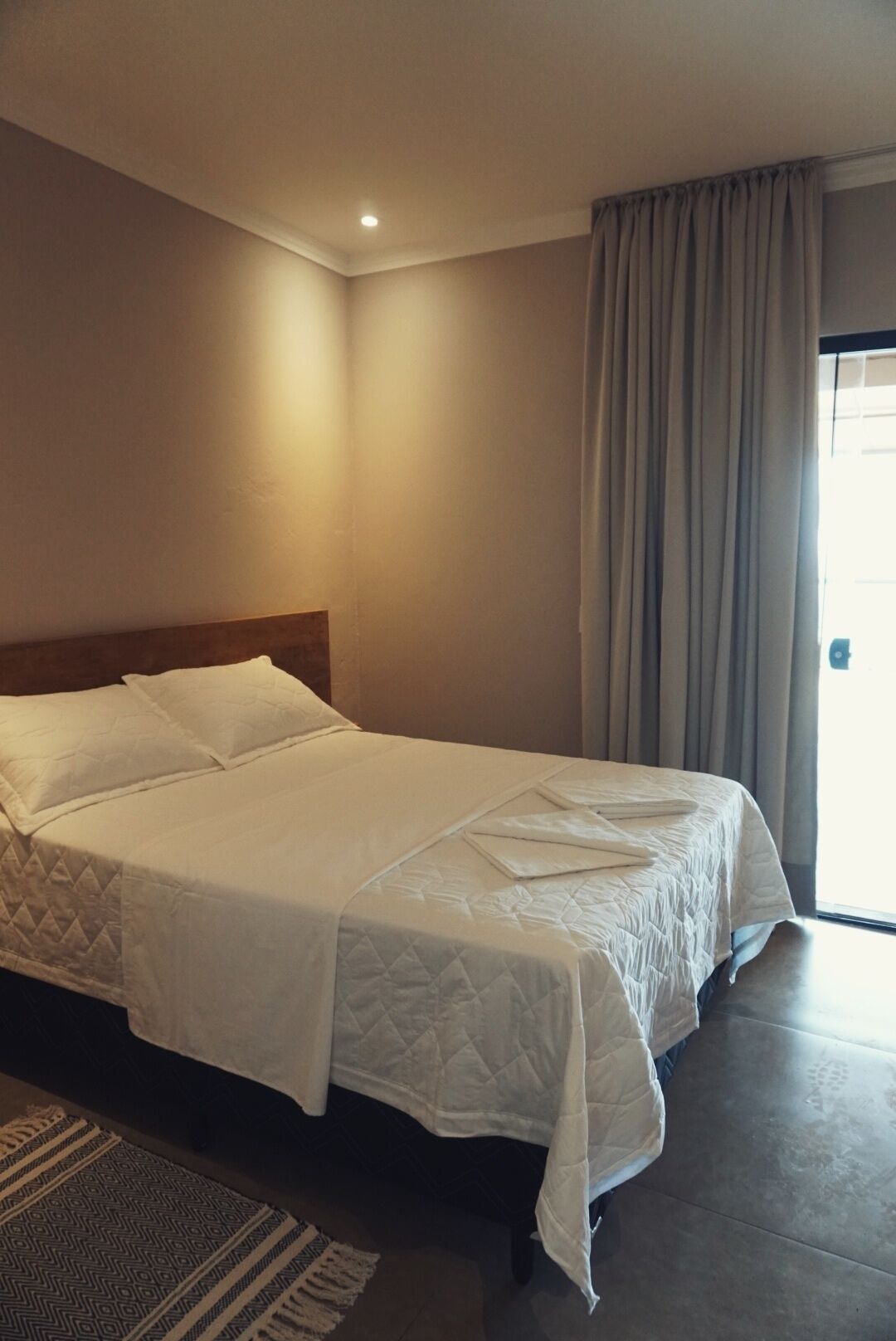 Quarto de hotel com uma cama grande, cabeceira de madeira, janela com cortinas e um tapete pequeno.