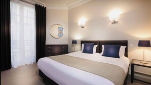 A hotel room with a large bed, two bedside tables with lamps, a round mirror, and a window with curtains.