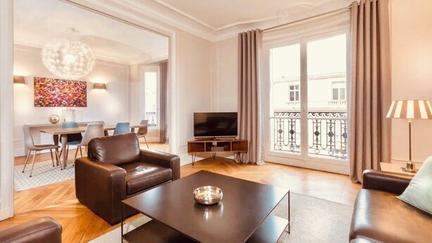 A modern living room with leather sofas, a coffee table, a TV, and a balcony with curtains.