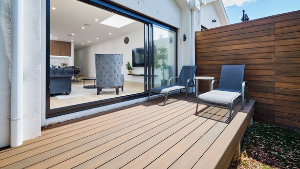 Eine moderne Außenterrasse mit Holzboden, einer Glastür, die zu einem Innenbereich führt, und zwei Lounge-Stühlen.