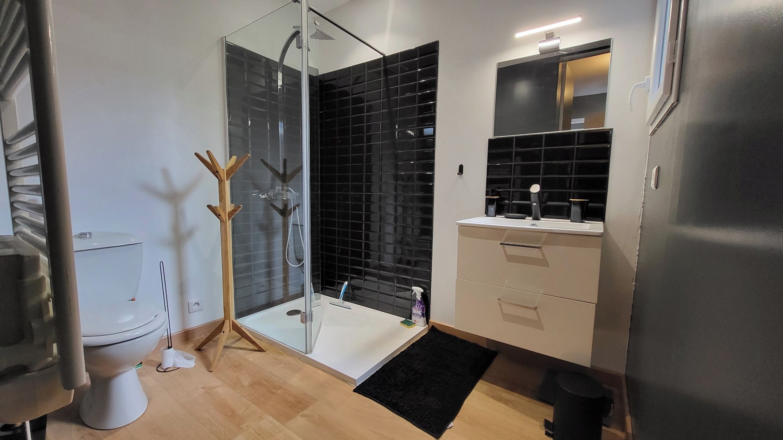 A modern bathroom with a glass shower enclosure, a white toilet, a beige vanity with a mirror, and a black rug.