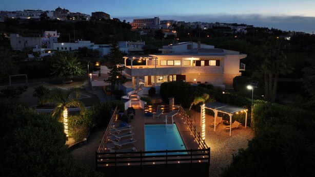 Ein modernes, zweistöckiges Haus mit Swimmingpool, umgeben von angelegten Gärten und Blick auf eine Hügelstadt.