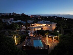 A modern two-story house with a swimming pool, surrounded by landscaped gardens and a view of a hillside town.
