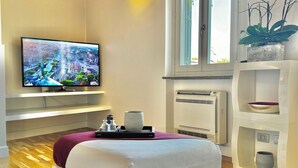 A modern hotel room with a flat-screen TV, a bed, a white console table, a potted plant, and a window with a view of the city. - Vetrina Roma - Stay in Luxury (Roma)