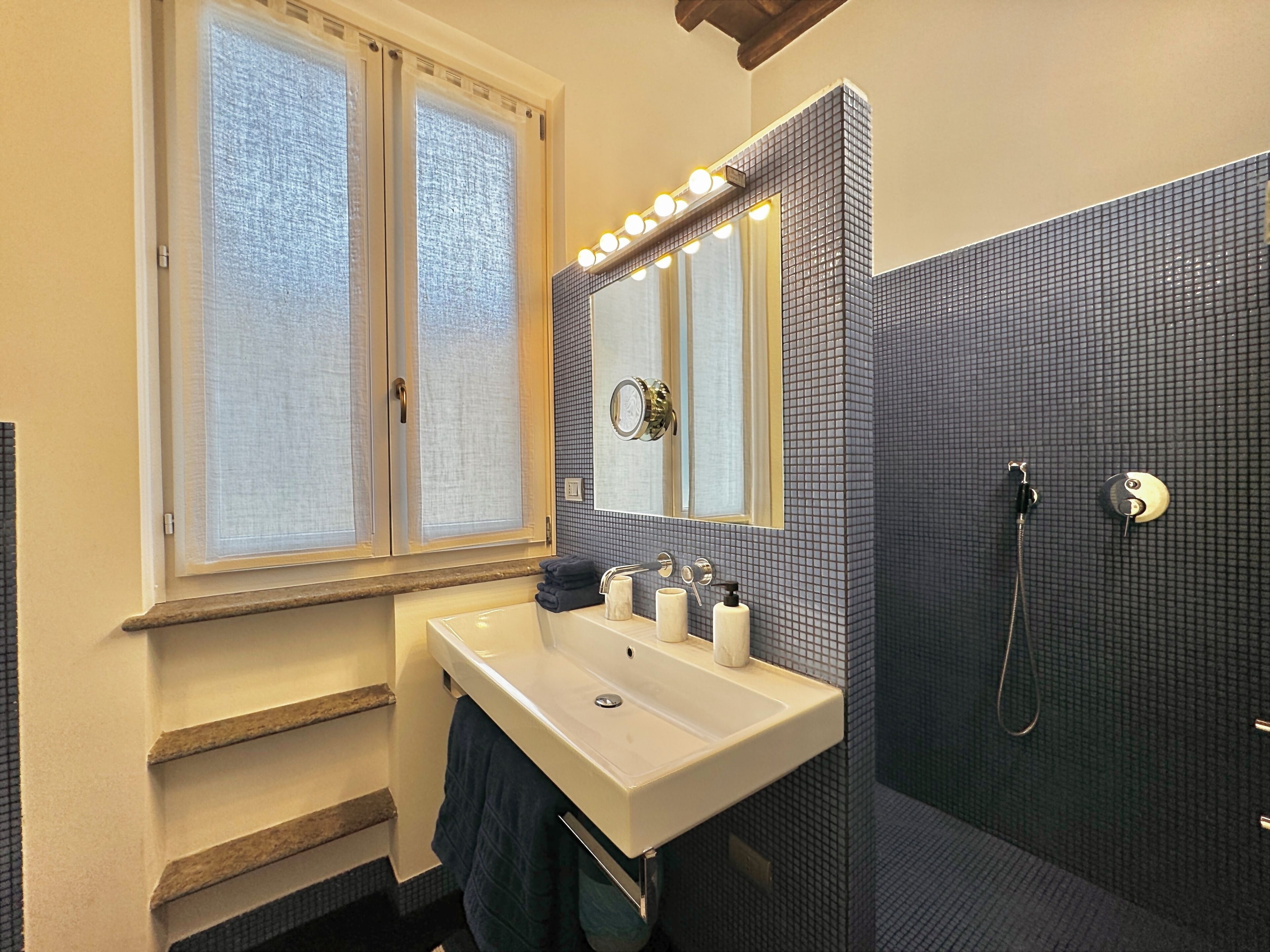 A bathroom with a white sink, a mirror with lights, a shower, and a window with blinds.