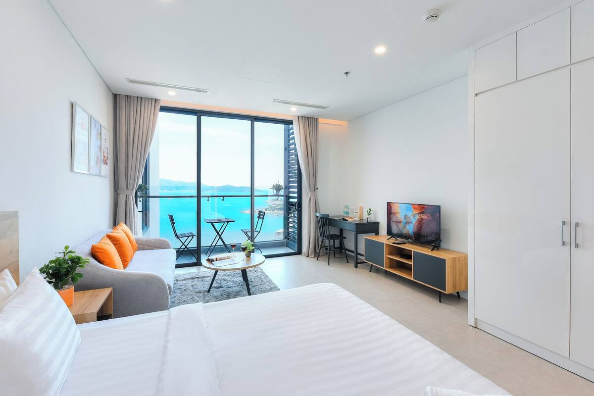 A modern hotel room with a large bed, a sofa, a dining area, and a TV. The room has a balcony with a view of the sea.