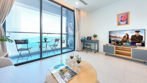 Un salon moderne doté d’une grande porte-fenêtre coulissante donnant sur un balcon avec vue sur la mer. On y trouve un téléviseur à écran plat posé sur un meuble, une table basse avec un magazine et des verres de vin, ainsi qu’un petit bureau avec un ordinateur portable.
