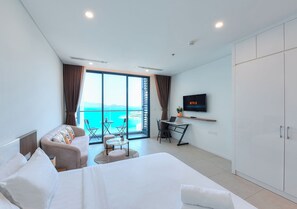 A modern hotel room with a large bed, a sofa, a desk with a laptop, a TV, and a balcony with a view of the sea.