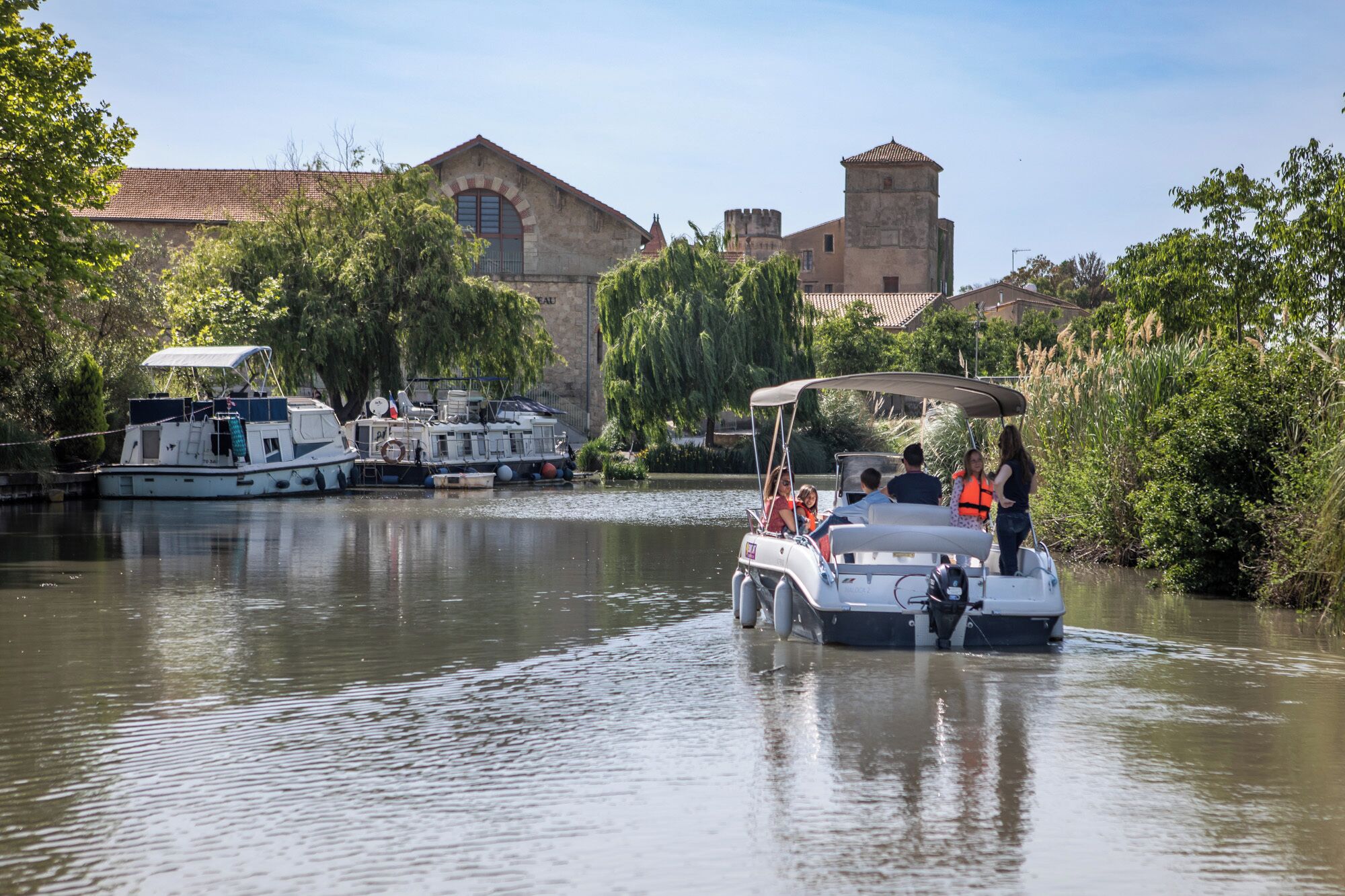 Een boot met passagiers op een kalme rivier, omringd door historische gebouwen en weelderige begroeiing.