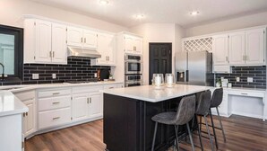 A modern kitchen with white cabinetry, a black backsplash, and a central island with bar stools. - Executive Rental 5BR 5BA, Arcade, Bounce House, GYM, Grill (Omaha)