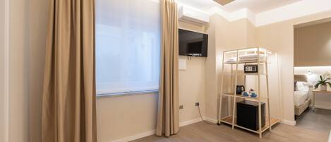 A modern hotel room with a large window, a flat-screen TV, a built-in mini-fridge, a bedside table with a lamp, and a bed with a headboard.