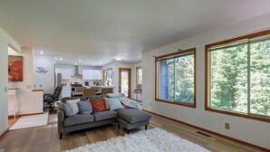 Un salon spacieux avec un canapé d’angle gris, un tapis blanc à poils longs et de grandes fenêtres donnant sur la forêt.