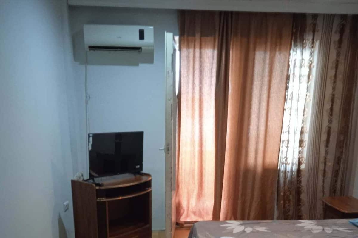 A hotel room with a bed, a TV on a wooden stand, a small table, and a window with curtains.