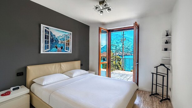 A bedroom with a bed, a nightstand, a bedside table, a window with a view, and a door leading to an outdoor area. - Casa A&E - Panoramic Lake Orta Views (Prà della Marta)