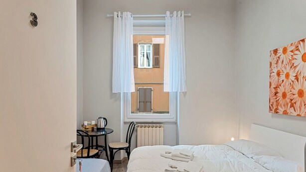 Ett hotellrum med en säng, ett bord med en vattenkokare, en stol och utsikt över omgivningen genom ett fönster med vita gardiner.