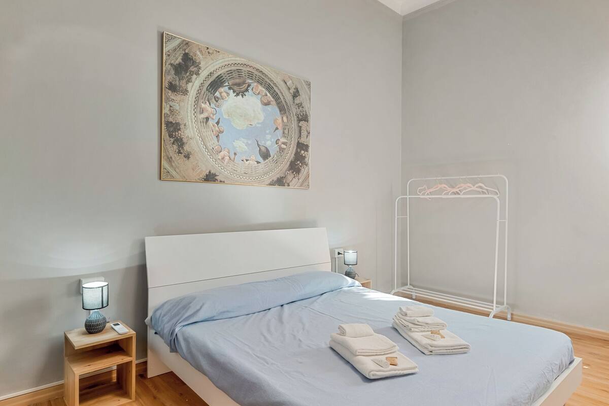 Un dormitorio con una cama, una mesita de noche con lámpara, una obra de arte en la pared y un perchero para toallas.