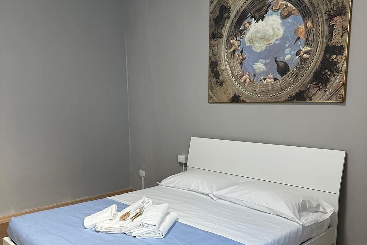 Una cama bien hecha con cubrecama azul y almohadas blancas, una toalla doblada y una obra de arte enmarcada encima.