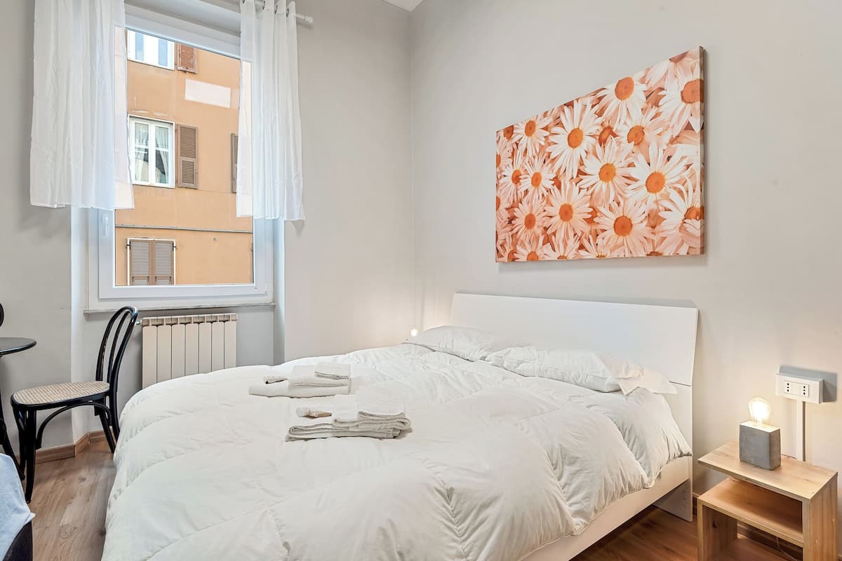 Una cama bien hecha con sábanas blancas, una mesita de noche con lámpara y una ventana con cortinas blancas.
