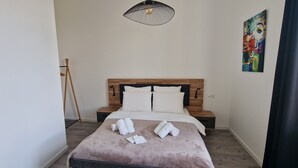 Een slaapkamer met een houten hoofdbord, wit beddengoed, een staande lamp en een kleurrijk schilderij aan de muur.