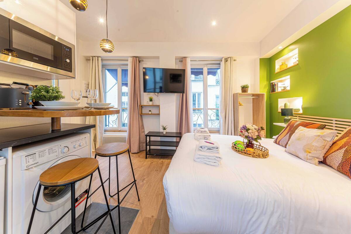 Habitación de hotel compacta con kitchenette, cama con ropa de cama blanca, televisor de pantalla plana y pared de acento verde.