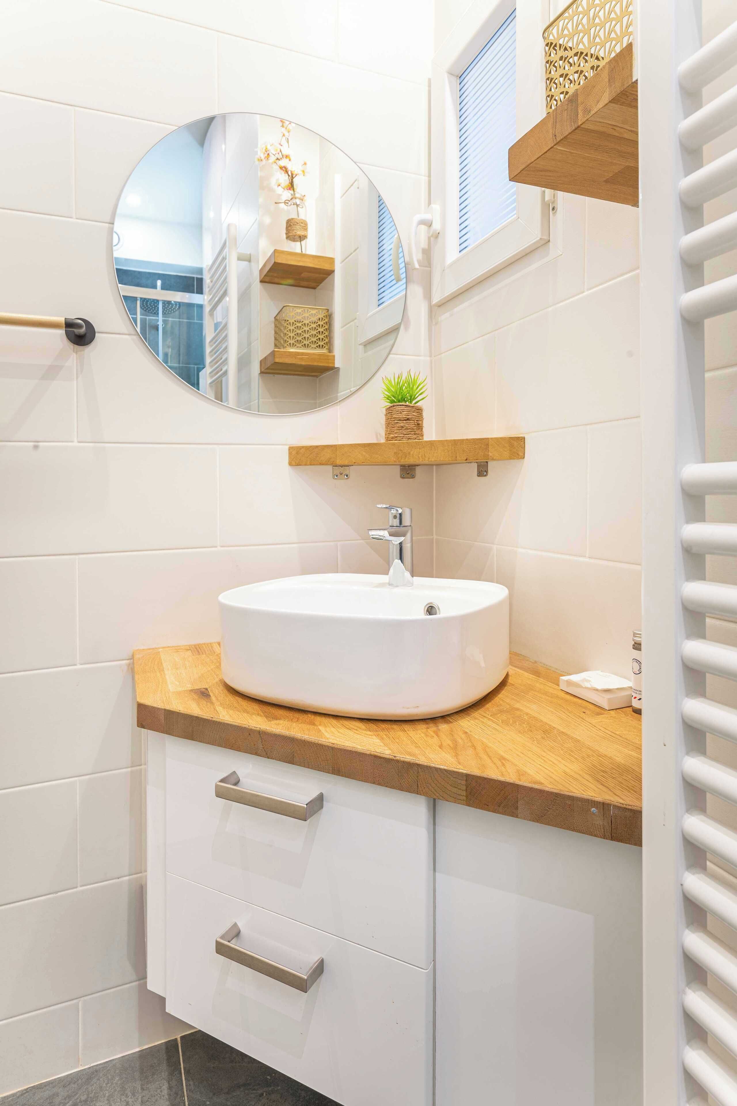A modern bathroom with a round mirror, a white sink, wooden shelves, and a towel rack.