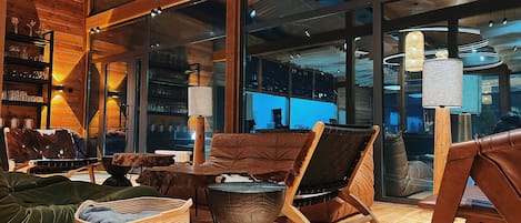 A cozy interior with wooden furniture, a fireplace, and large windows offering views of the outside.