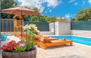 Un jardin avec une piscine, des chaises longues en bois, une structure de jeux en bois et des plantes en pot.
