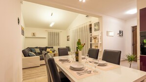 A dining area with a long table set for four, surrounded by chairs, with a sofa and a small plant in the background.
