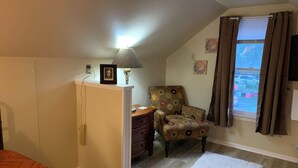 Quarto com cama, cômoda, cadeira, luminária, janela com cortinas e tapete.
