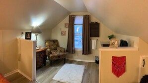 Um quarto de sótão pequeno e acolhedor, com teto inclinado, cômoda de madeira, poltrona estampada com flores, tapete branco, lenço vermelho e uma janela com cortinas.