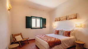 A bedroom with a bed, a chair, two side tables, a window with shutters, and wall-mounted lights.