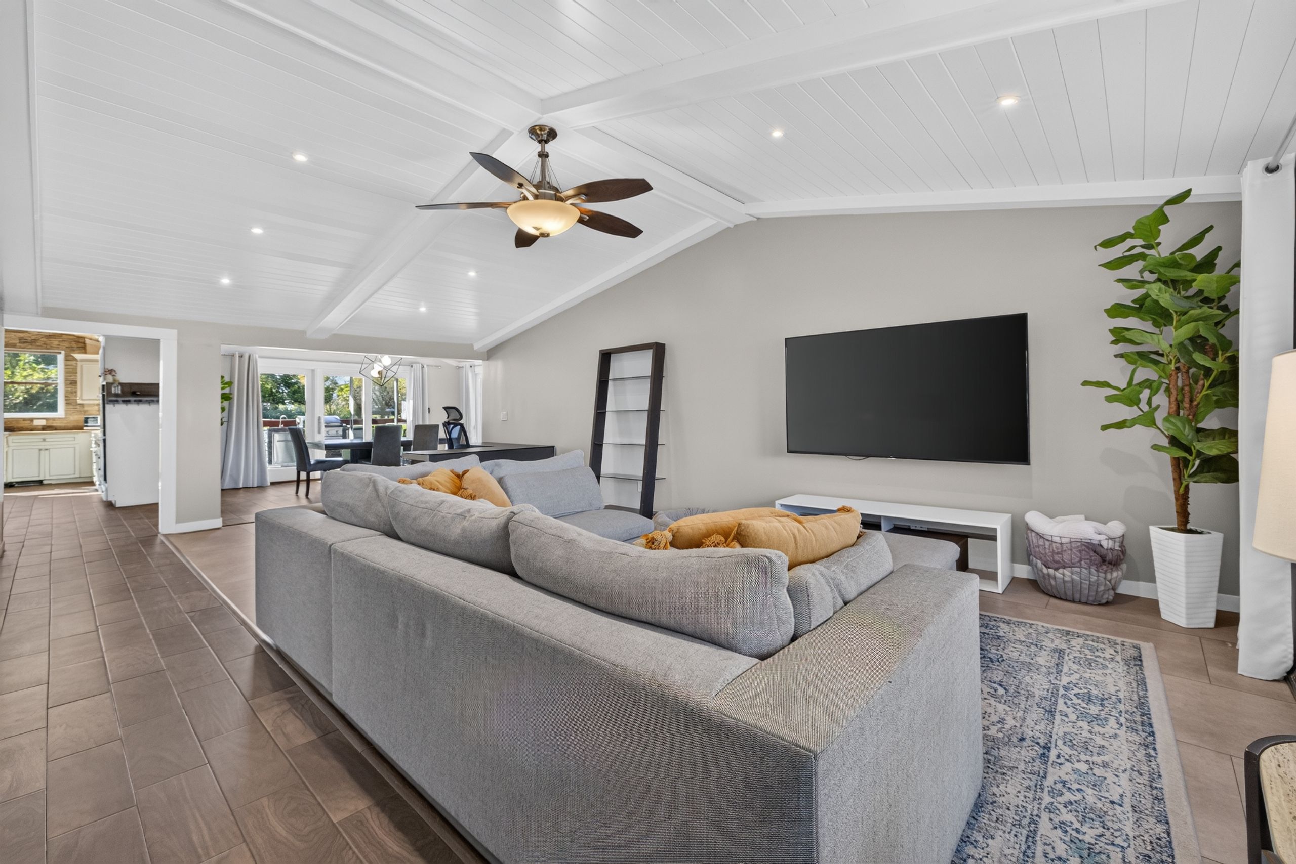 Un salon moderne avec un canapé d’angle gris, un téléviseur à écran plat fixé au mur et un ventilateur de plafond.
