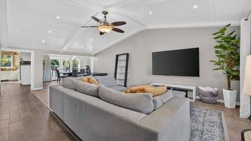 Un salon moderne avec un canapé d’angle gris, un téléviseur à écran plat fixé au mur et un ventilateur de plafond.