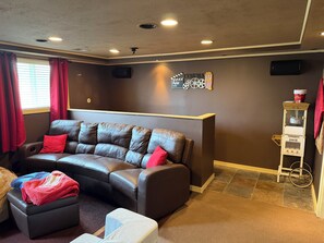 A living room with a leather sofa, a small ottoman, a red blanket, a cinema-themed wall decoration, and a candy dispenser.