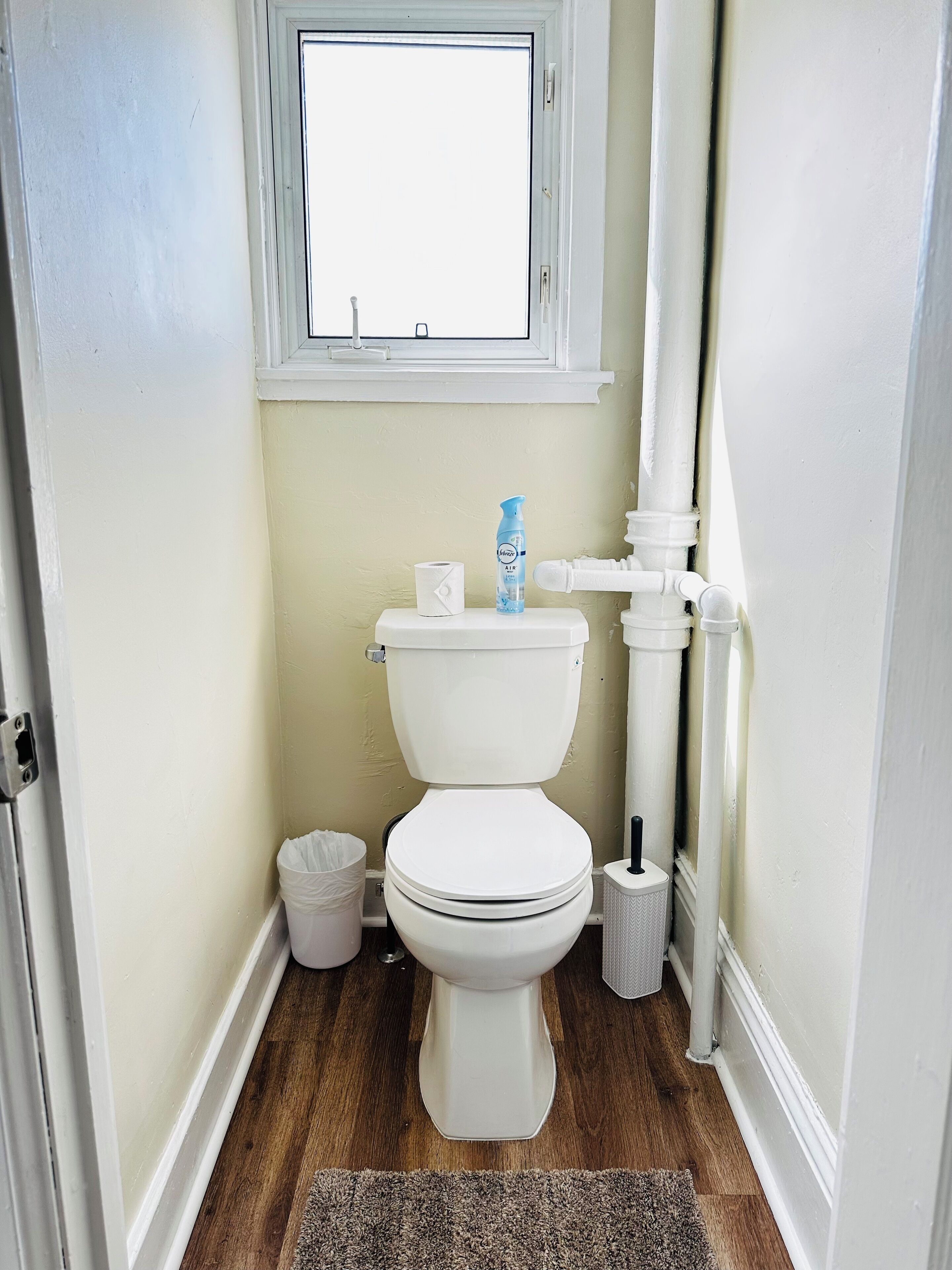 A small, clean bathroom with a toilet, a window, and a white trash bin.