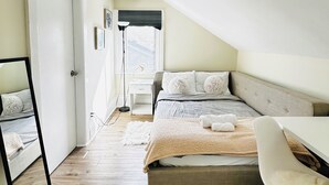 A bedroom with a sloped ceiling, a bed with a grey headboard, a bedside table, a chair, and a window with a view. - Spacious 2BR House with Netflix, BBQ, and Patio Near City Centre (Peterborough)