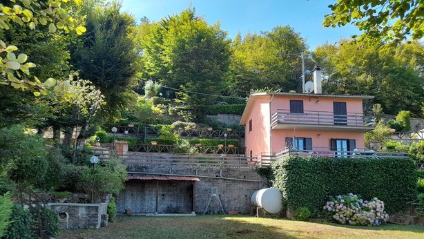 Une maison à deux étages dotée d’un balcon, entourée d’une végétation luxuriante et d’un mur de pierre.