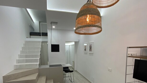 A modern living room with a staircase, a round white ottoman, and a TV on a metal stand.