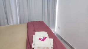 Een eenpersoonsbed met een rode spreideken en een witte deken, een klein wit kussen, en een roze en witte handdoek op het bed.