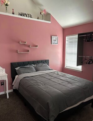 Une chambre à coucher comprenant un lit, une table de chevet, une étagère avec des objets décoratifs, une fenêtre avec des stores et un porte-manteau fixé au mur.
