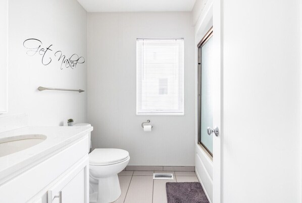 A modern bathroom with a white sink, toilet, and shower. A towel rack and a small potted plant are visible.
