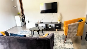 A modern living room with a dark sofa, a small round table, a yellow armchair, a flat-screen TV mounted on the wall, and a washing machine in the corner. - Yo House (Kampala)