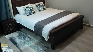 A neatly made bed with a headboard, a bedside table with drawers, a wall-mounted light, and a framed picture on the wall. - Yo House (Kampala)