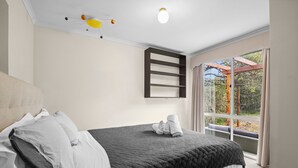 A bedroom with a bed, pillows, a quilted bedspread, a ceiling light fixture, and a window with a view of trees. - Walk2Everything (Anglesea)