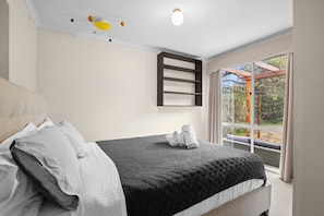 A bedroom with a bed, pillows, a quilted bedspread, a ceiling light fixture, and a window with a view of trees. - Walk2Everything (Anglesea)