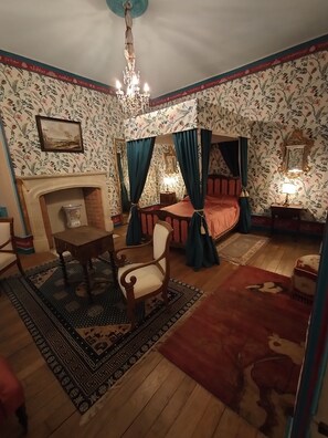 Quarto tradicional com cama de dossel, lareira, candelabro e papel de parede estampado.