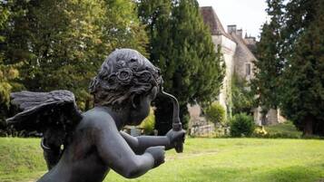 Een standbeeld van een figuur met vleugels en een trompet, gelegen in een grasrijk gebied met bomen en een gebouw op de achtergrond.