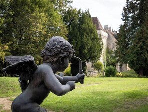Uma estátua de uma figura alada segurando uma trombeta, em uma área gramada com árvores e um edifício ao fundo.